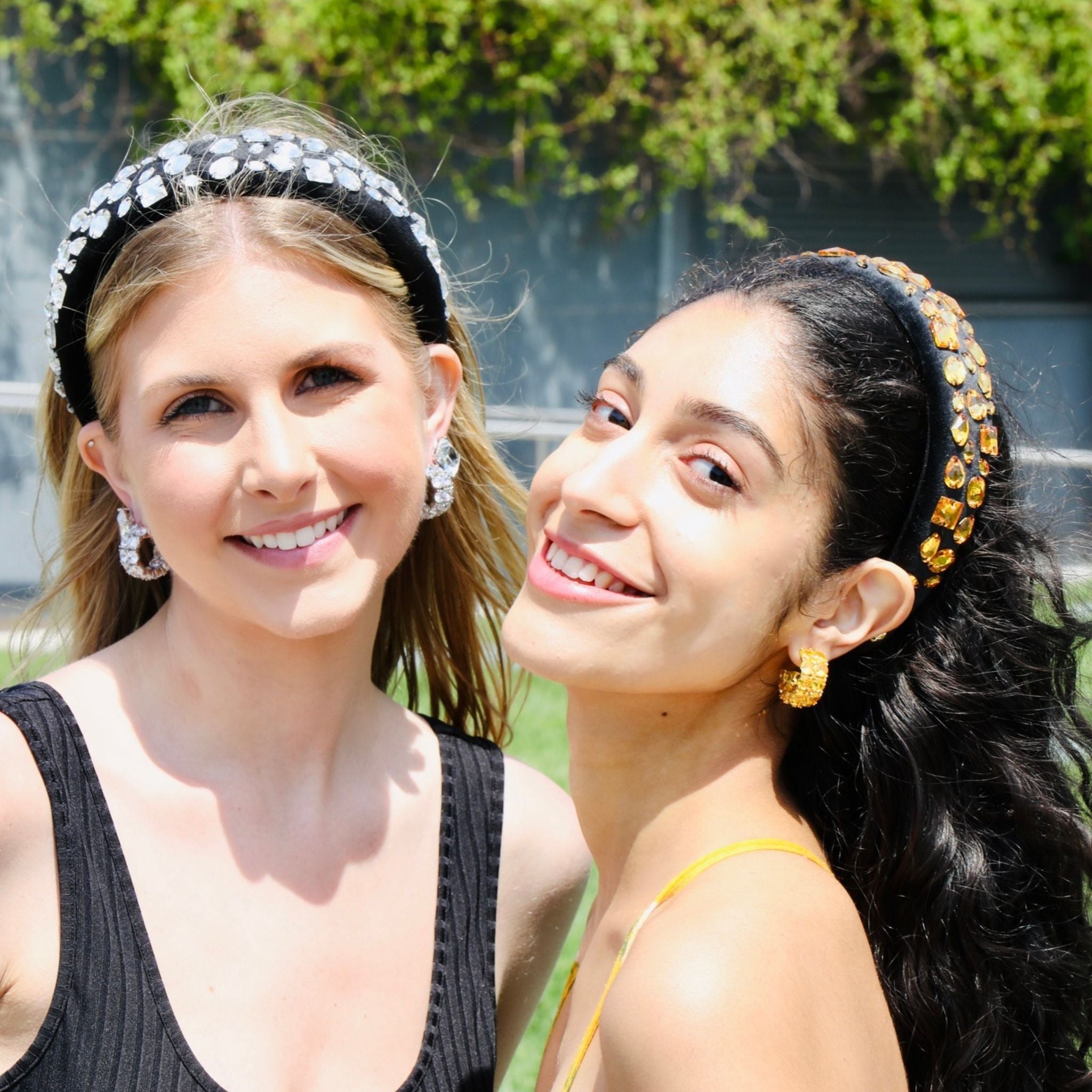 Two women smiling outdoors, each wearing a decorative embellished black headband. The one the left is silver-like rhinestone headband, while the headband on the right is yellow stone.